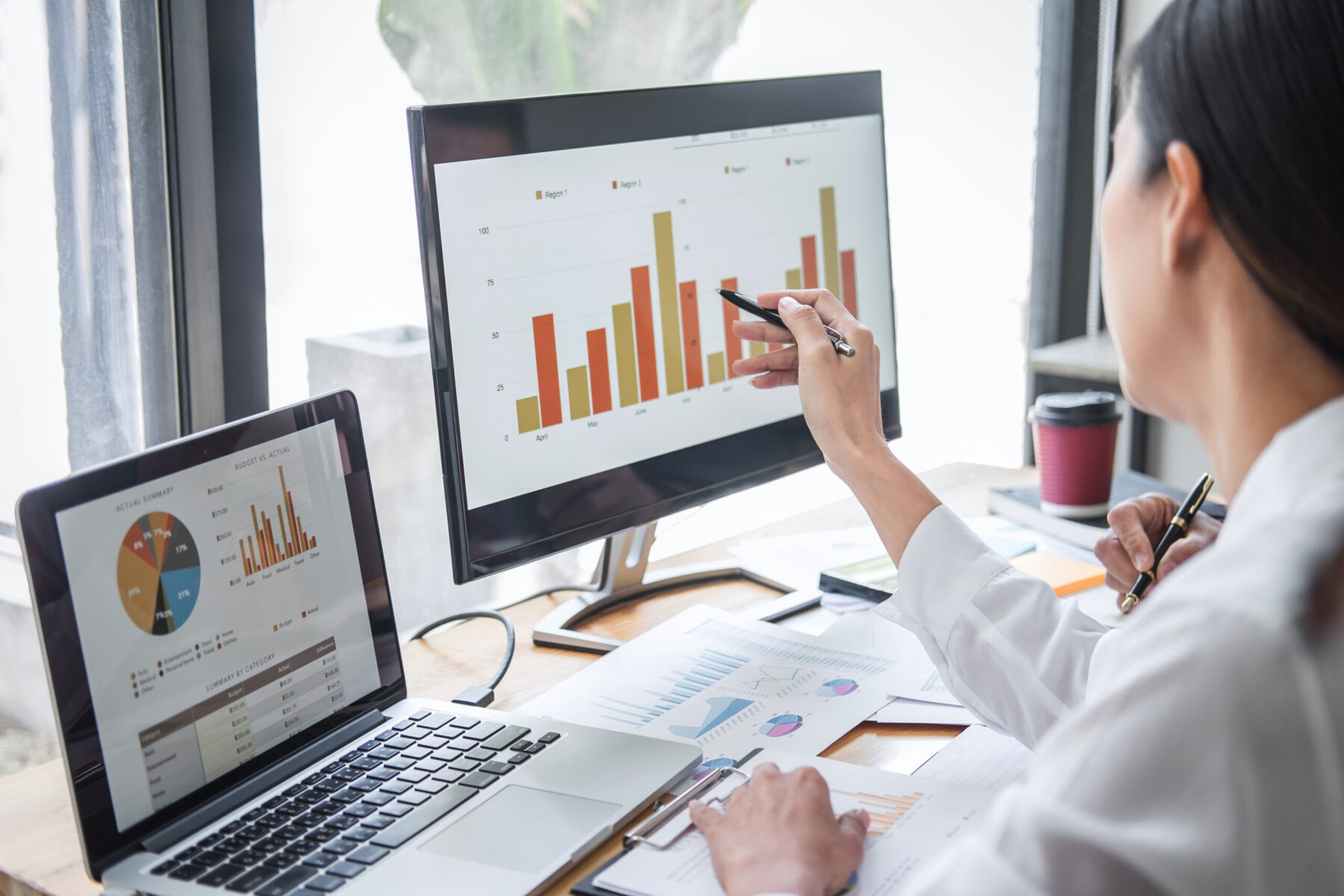 Two professional financial analysts review performance indicator charts on a laptop and second monitor in a modern office.