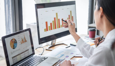 Two professional financial analysts review performance indicator charts on a laptop and second monitor in a modern office.